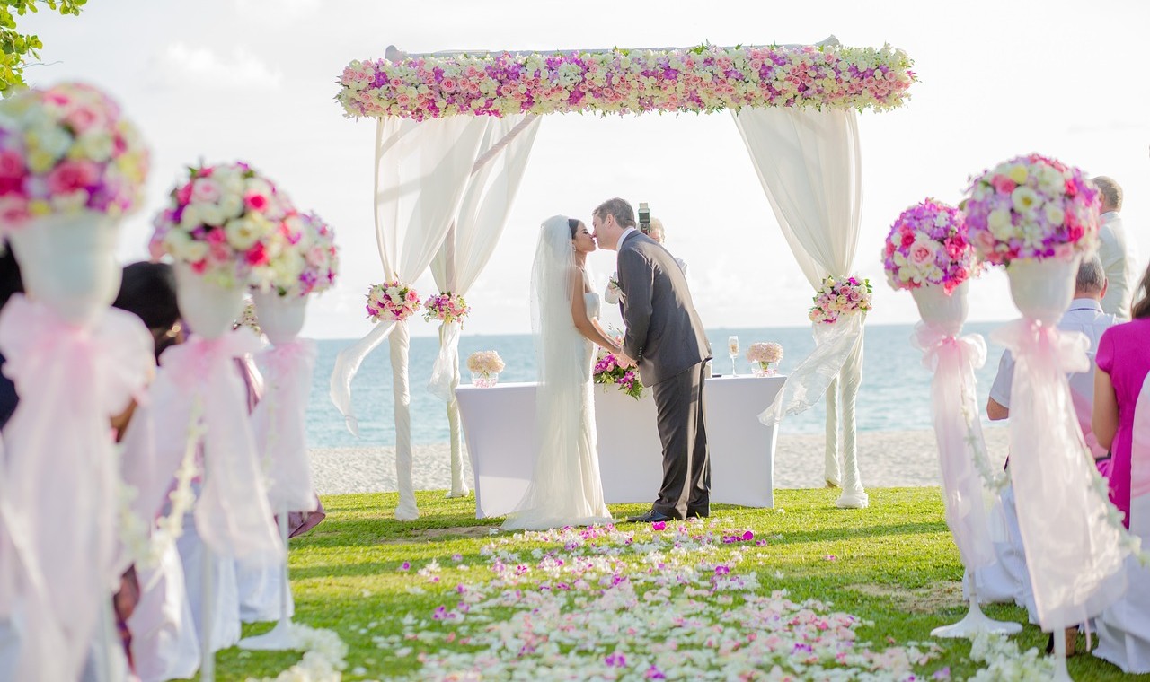 A bride and groom kiss under a flower arch at a beach wedding while their guests look on